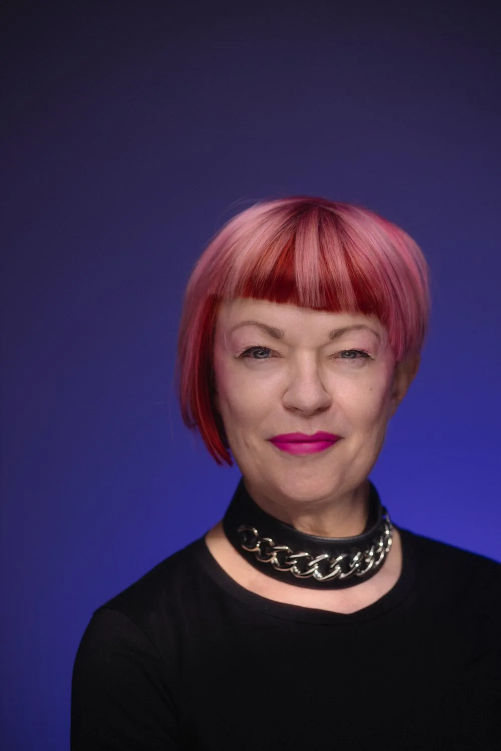Portrait of Vivienne Austin, with a sharp pink and red bob, magenta lipstick and a black chain-link choker